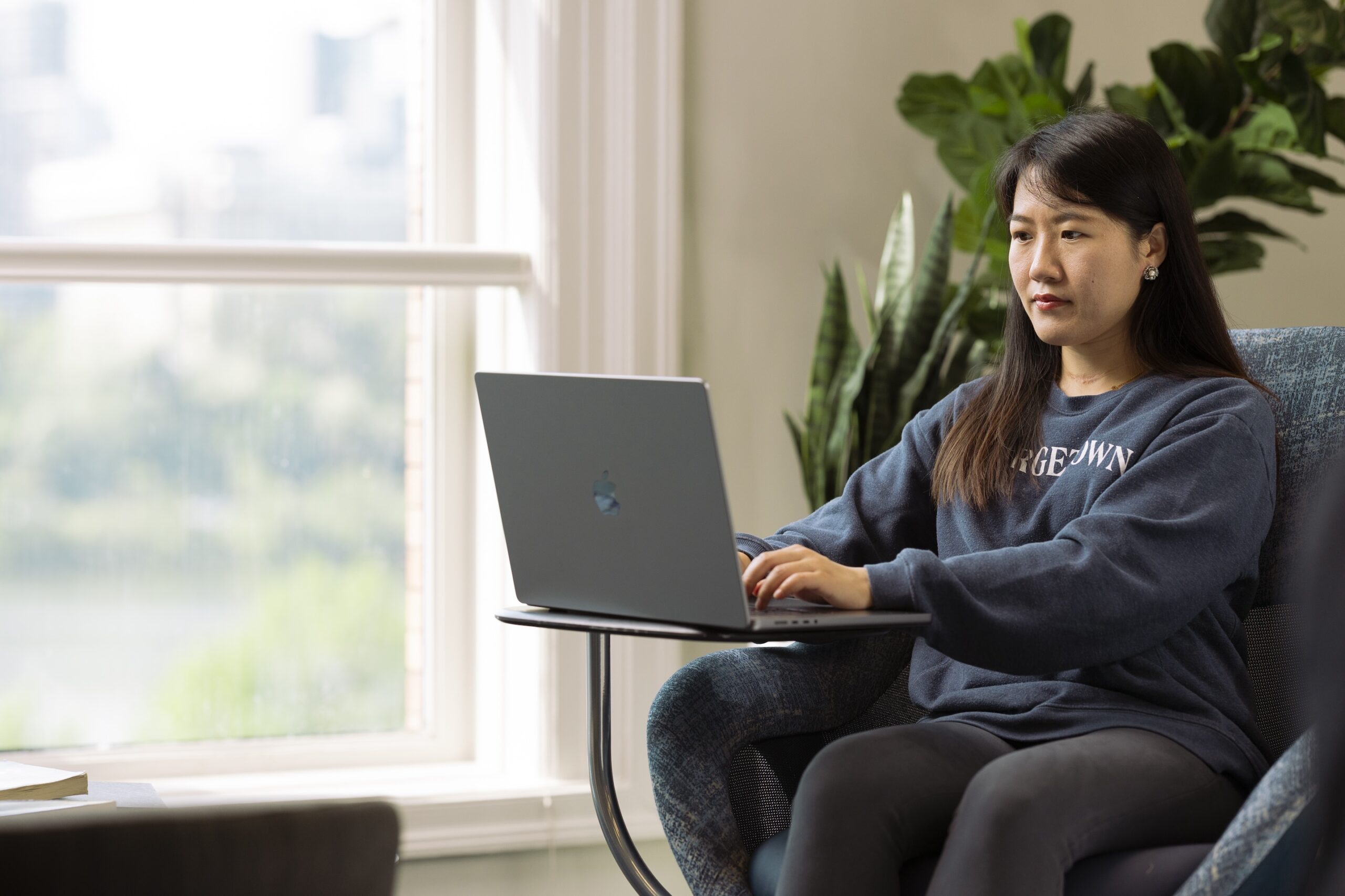 Woman wearing a navy blue Georgetown sweatshirt working on a laptop with a plant and a window in the background